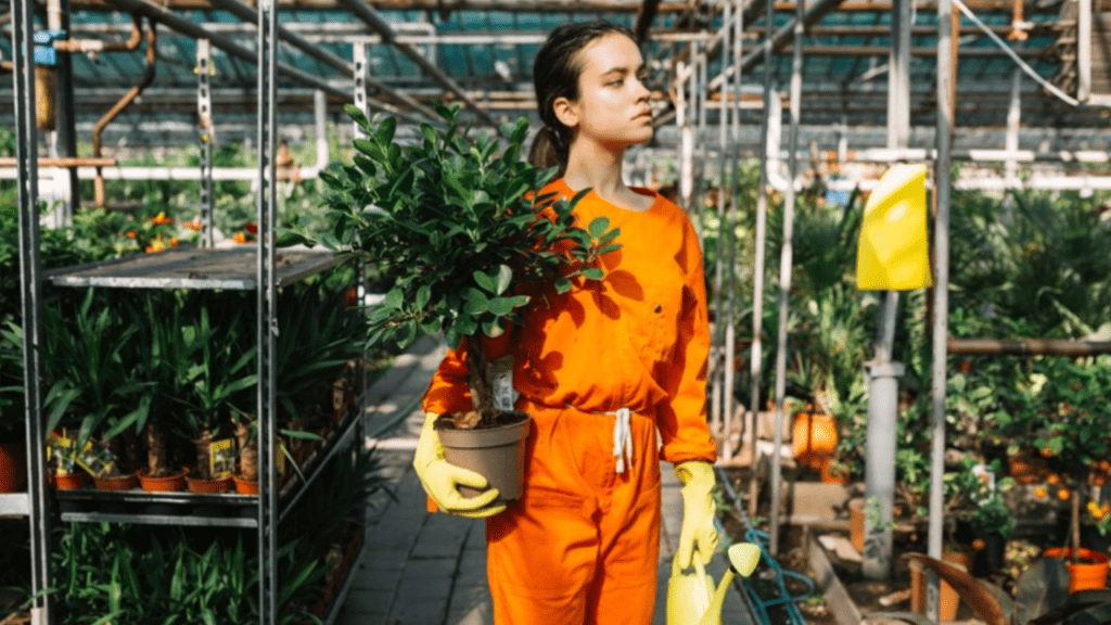 A woman in a nursery dressed in orange and wearing yellow globes holds a plant with a pot in one hand and a watering can in the other.