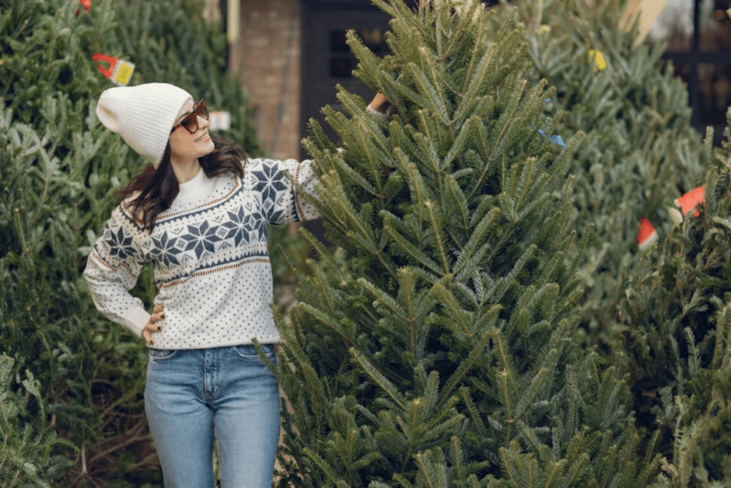 A woman wearing a cozy sweater and hat stands joyfully in front of a beautifully decorated Christmas tree.