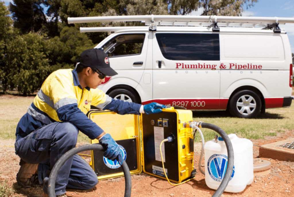 Worker in hi-vis gear operating equipment near a plumbing service van.