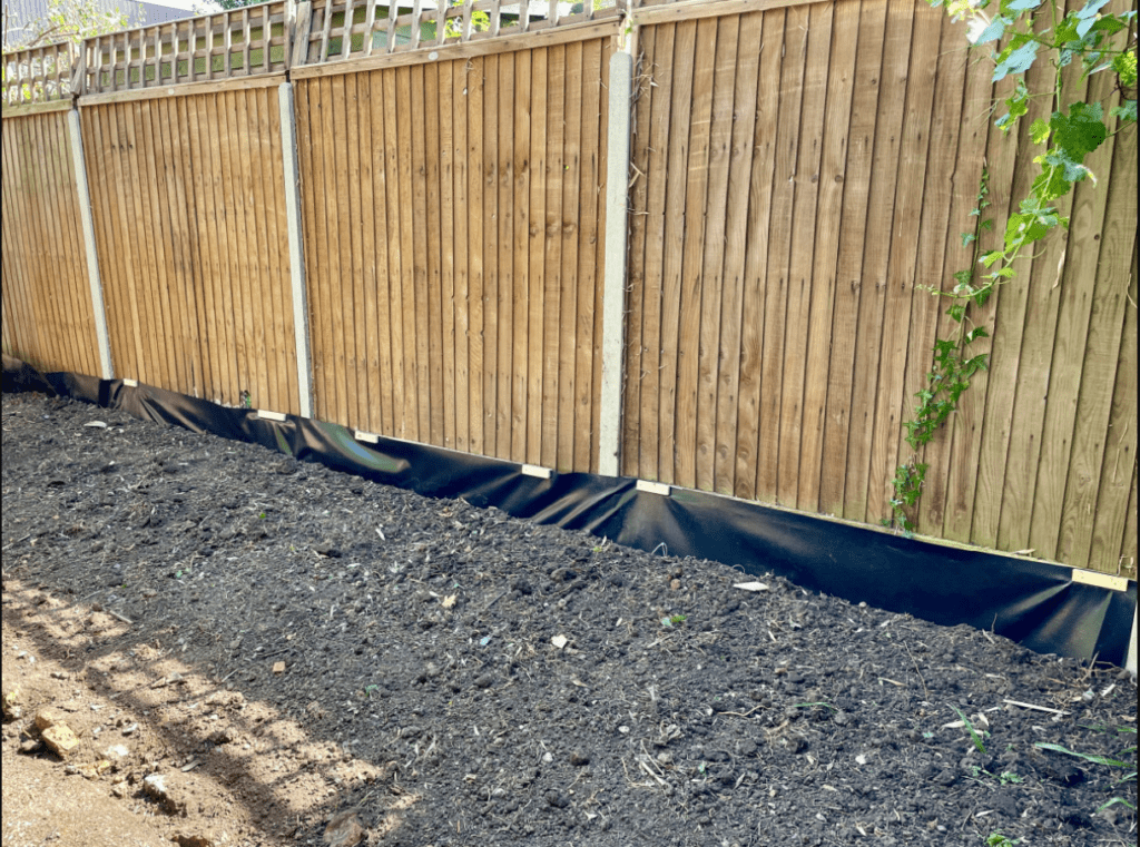 Wooden fence with landscape fabric at the bottom along a dirt patch with sparse vegetation.