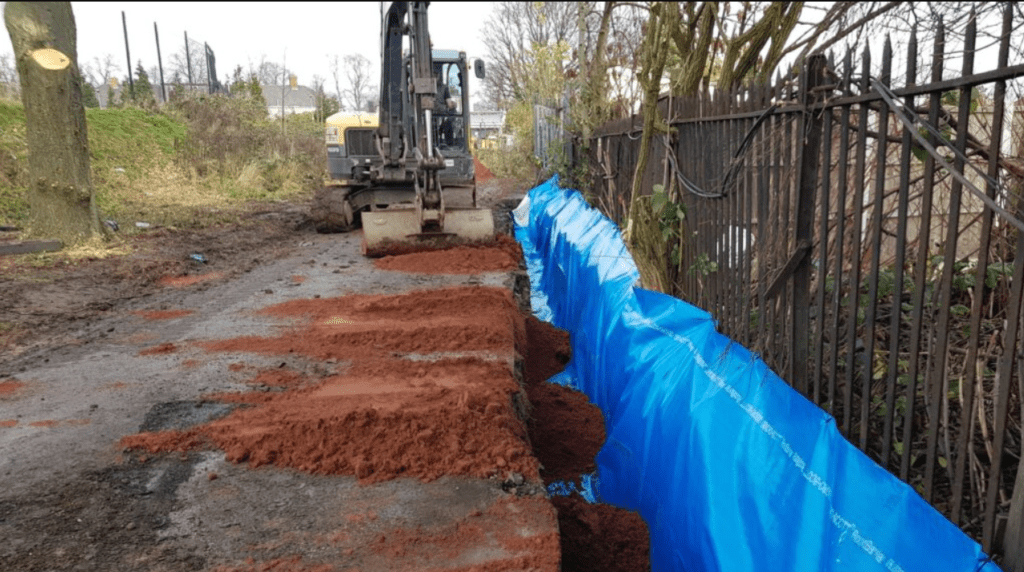 Excavator by a trench lined with blue tarps and fresh soil, near a metal fence.