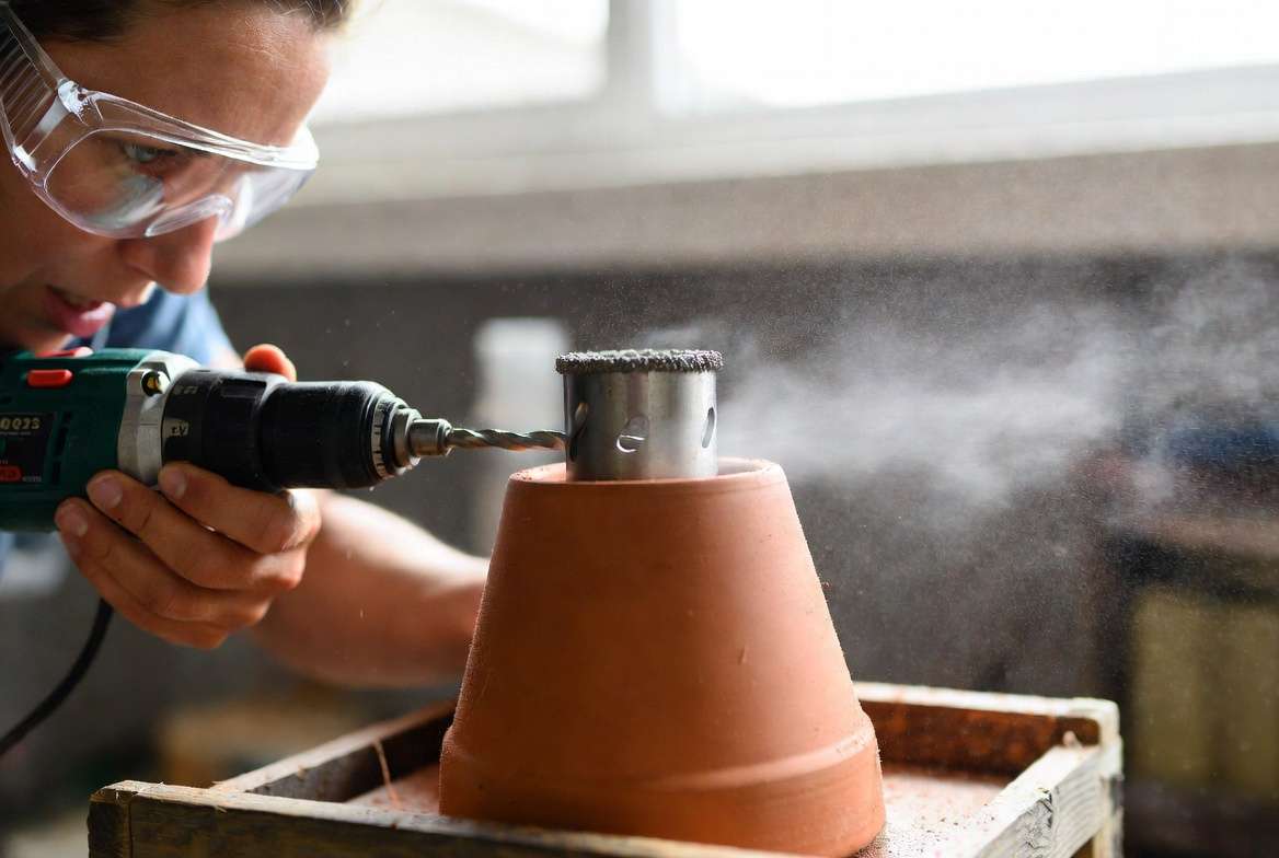 An expert using a power drill with a diamond hole saw bit to drill a new drainage hole into a ceramic planter.