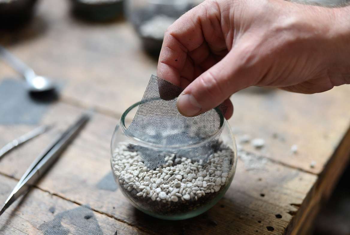 A gardener’s hand placing a mesh filtration barrier over pebbles inside a miniature glass terrarium to prevent soil movement.