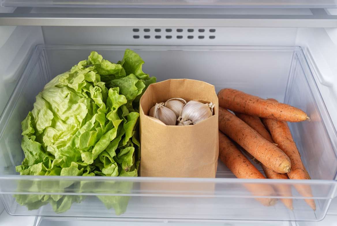 A brown paper bag of garlic bulbs chilling inside a refrigerator crisper drawer with other vegetables.